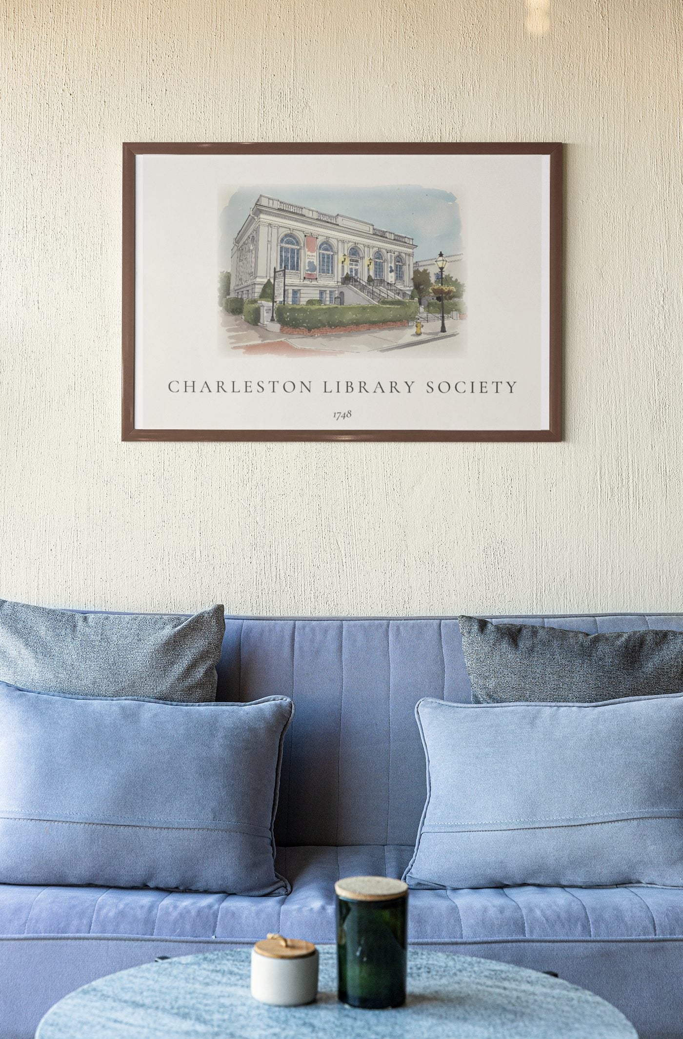 An ink and wash illustration of the Charleston Library Society, framed on a textured beige wall above a soft blue sofa.