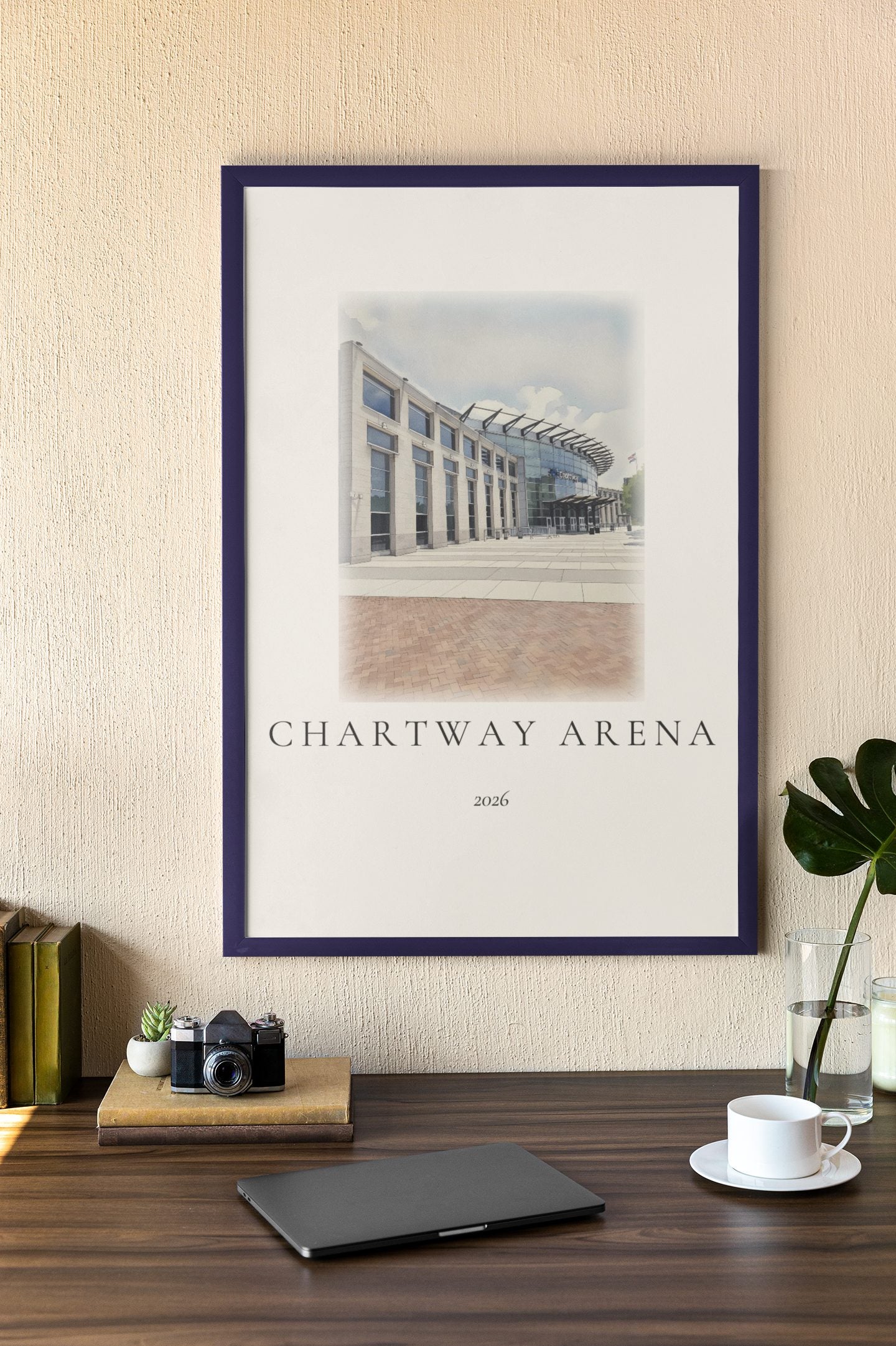 An ink and wash illustration of Chartway Arena framed on an office wall, with a camera and coffee on a wooden desk.